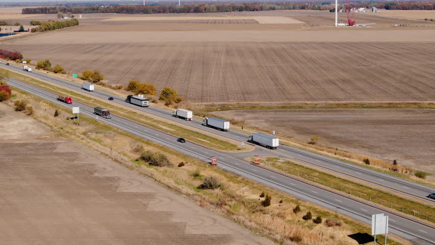 Multiple semi-trucks and cars traveling along a busy highway surrounded by flat plowed agricultural fields. Long-haul transportation, logistics, and supply chain