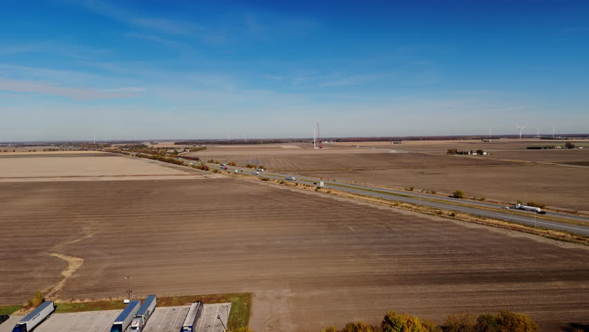 Multiple semi-trucks and cars traveling along a busy highway surrounded by flat plowed agricultural fields. Shoot of long-haul transportation, logistics, and supply chain