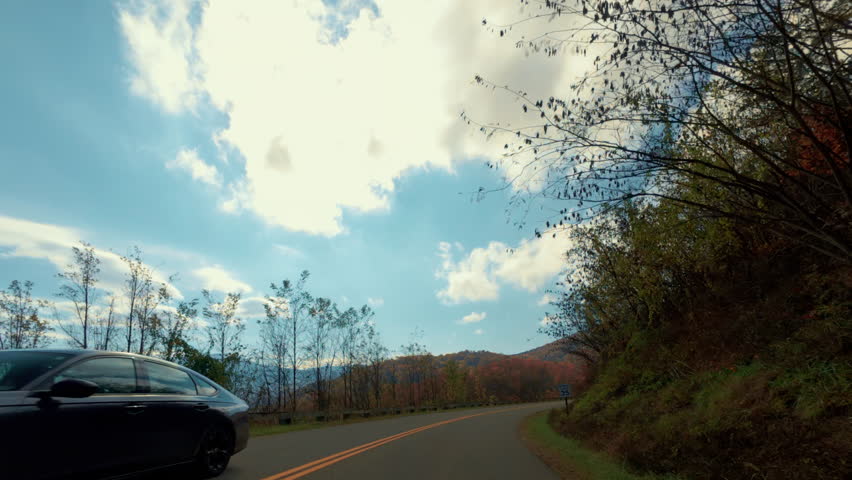 Driving through a vibrant autumn forest, with colorful fall foliage surrounding a winding mountain road way. Tall trees in shades of red, orange, and yellow line roadway. 