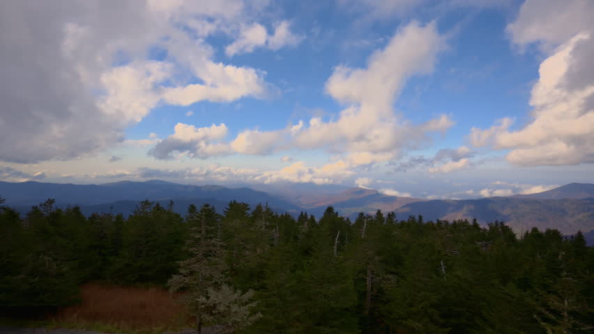 Aerial view of a mountain landscape with dramatic clouds drifting across blue sky. Rolling ridges stretch into the distance, illuminated by soft light. Peaceful wilderness