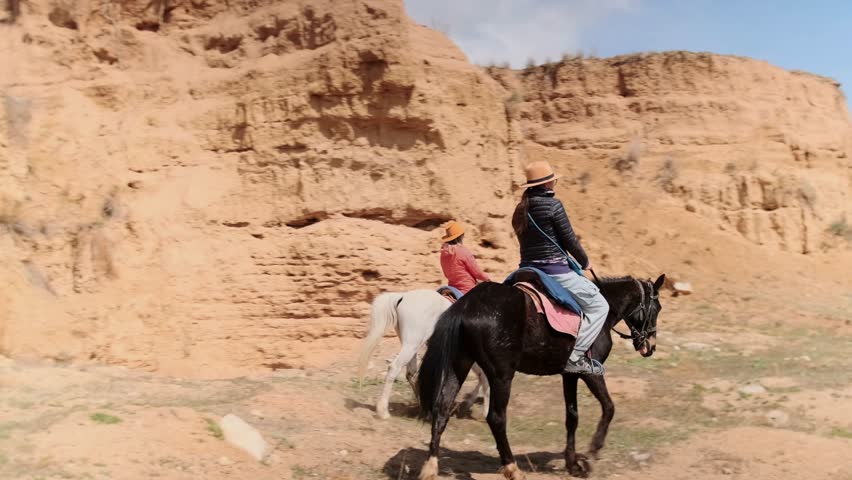 Two women in hats ride horseback through a dramatic canyon landscape on a clear, sunny day. The riders, wearing casual travel clothes, move slowly past orange sandstone cliffs. 
