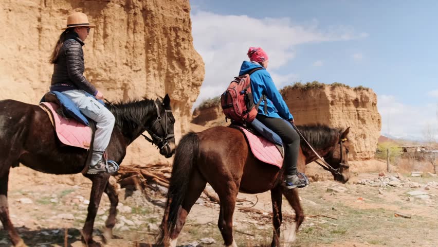 Two riders, travels across open canyon terrain on horseback. Women ride at ease through wide, wild nature, surrounded by dry cliffs and open skies, freedom and escape from routine. Side view.