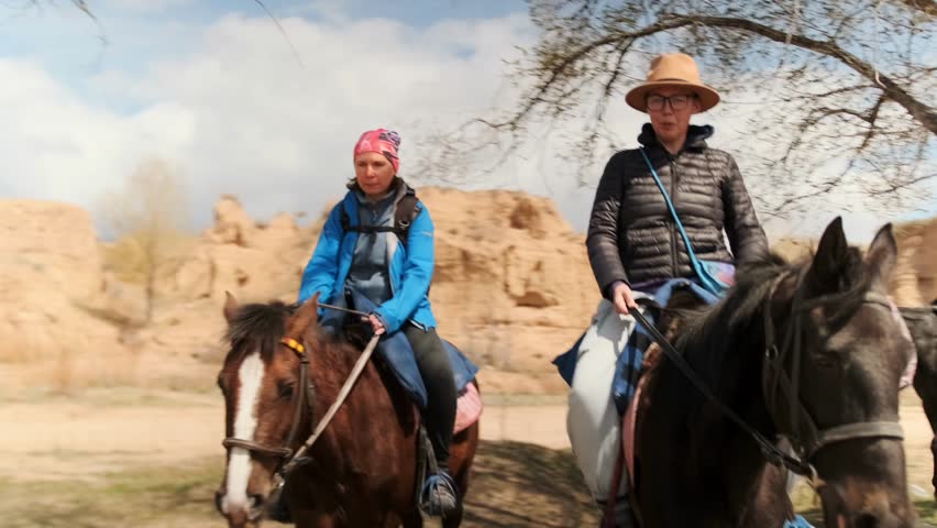 candid horse riders with scenic background, two relaxed riders on horses beneath scenic trees, pair of riders on horseback posing naturally amidst scenic desert and cliff scenery under bright daylight