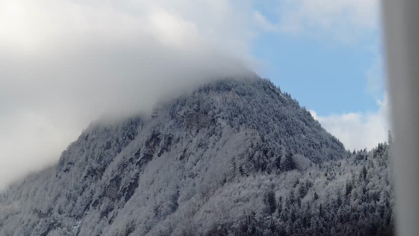 Snow-covered mountain with its top wrapped in fog under a partially blue sky. The scene captures a calm alpine winter atmosphere with clouds meeting rugged slopes.