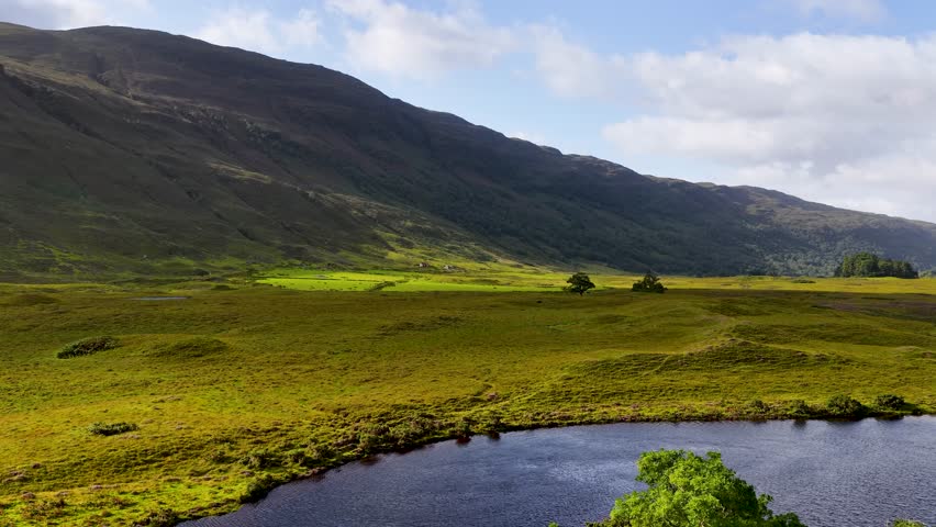 Drone pans across lush green meadow, reflective lake, and distant mountains under soft daylight