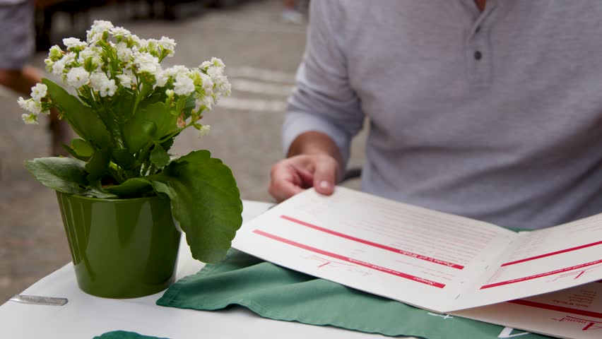 Man flips through menu at outdoor cafÃ© table with flowerpot, natural daylight, steady camera