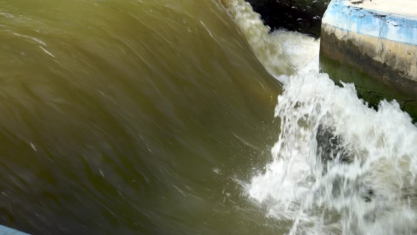 Powerful and rapid flow of brown, turbid water from a dam or sluice gate, creating intense white foam and spray.