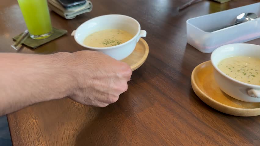 Hand sets down cream soup bowl on wooden table in cozy, naturally lit Bangkok cafe
