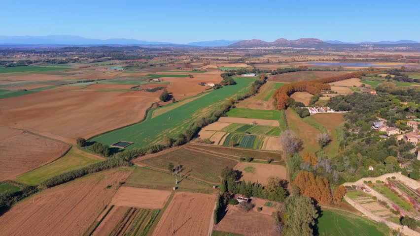 Breathtaking aerial footage showcasing the stunning patchwork of agricultural fields and autumn trees in the rural landscape of pals, costa brava, with distant mountains under a clear blue sky