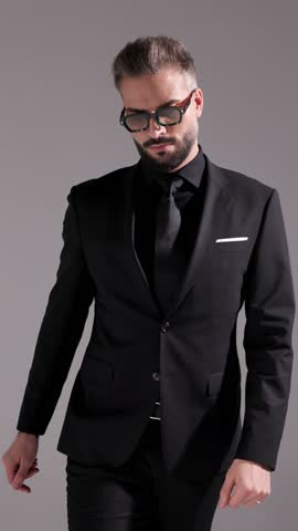 Serious man in sunglasses and all black suit is putting hands in pockets, fixing his lapel, crossing his arms, fixing his glasses, and walking away on grey background.