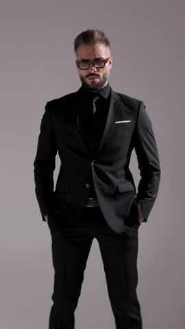 Cool man in sunglasses and all black suit is posing with hands in pockets, looking around, and walking away on grey background.