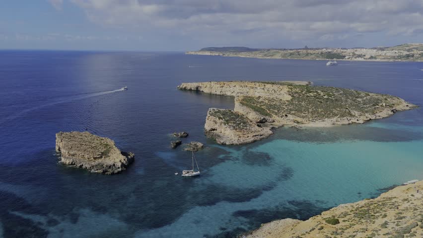 A high drone view over the Blue Lagoon shows Gozo, the open sea, part of Comino and Cominotto Beach with its cave, several yachts and a boat moving across calm turquoise water under sunny skies.