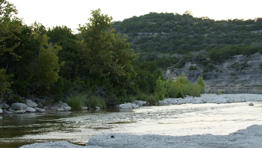 A peaceful riverside scene in a rocky, Central Texas landscape. A shallow river gently flows across limestone banks.