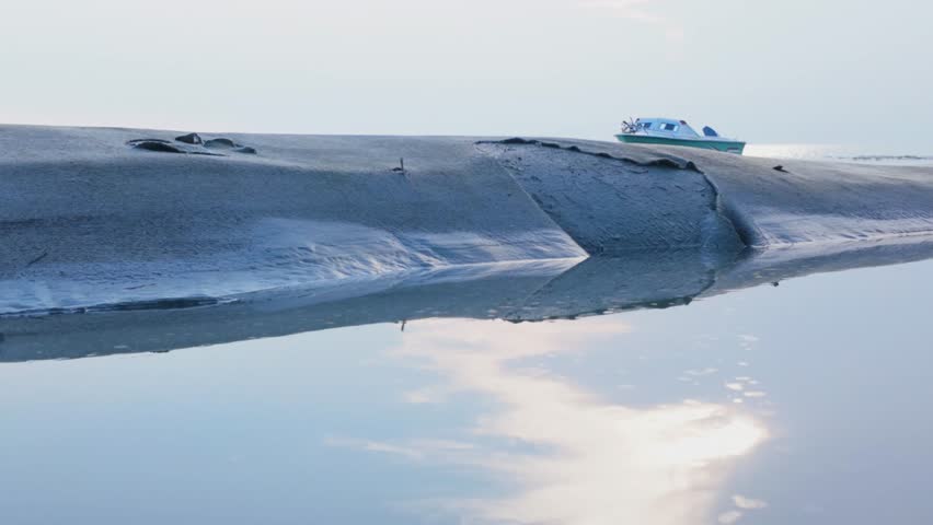 Serene coastal landscape featuring a smooth sandbank at low tide with clear water reflecting the sky. A small boat rests on the distant shoreline, creating a peaceful and minimal seaside atmosphere. 