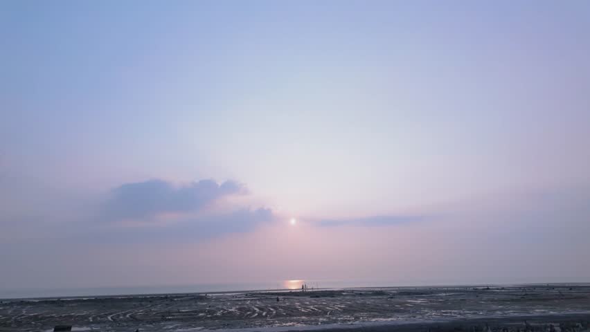 Serene coastal sunset scene featuring a soft pastel sky, gentle clouds, and the sun reflecting on the calm ocean horizon. The foreground shows a wide, textured shoreline at low tide.