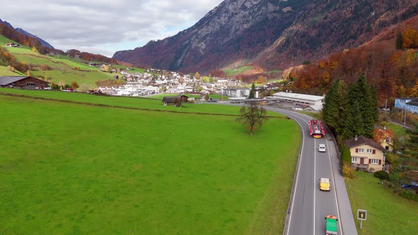 Aerial flyover driving cars and truck on street along river in Schwanden, Switzerland. Colored trees in fall season. green pasture with huts. City in distance.
