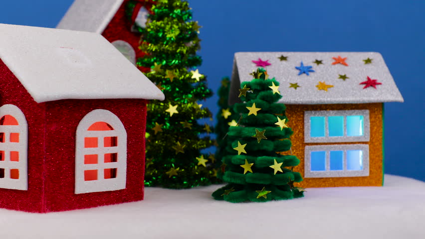 On a blue background, close-ups of rotating Christmas houses with bright lights in the windows and small Christmas trees
