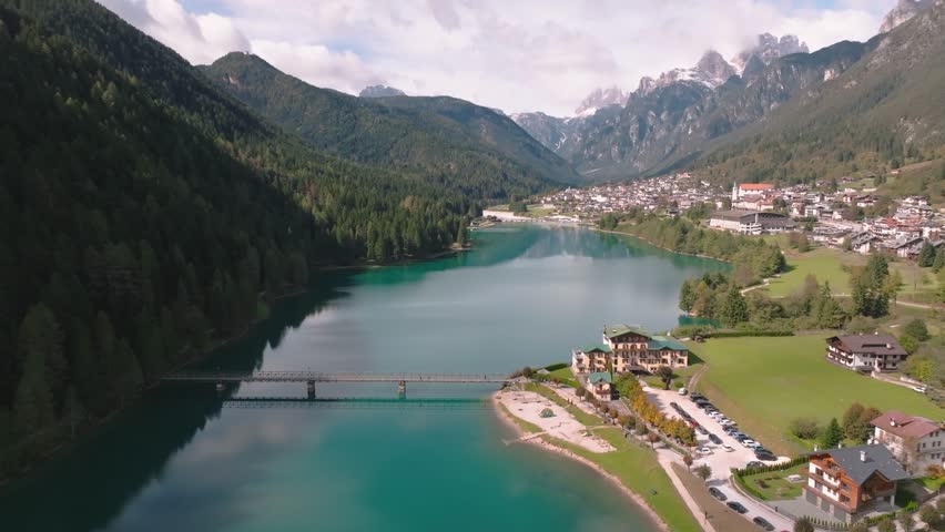 Aerial view of a bridge crossing vibrant turquoise water, leading to a building with a green roof, complemented by lush green grass, Misurina, Veneto, italy.