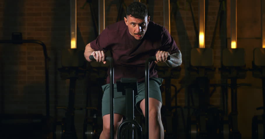 Slow Motion Of Athlete Training On Air Bike In Dark Gym With Dramatic Cinematic Lighting And Intense Shadows, Close Up Of Physical Effort And Sweat, Concept Of Endurance, Discipline, Inner Strength