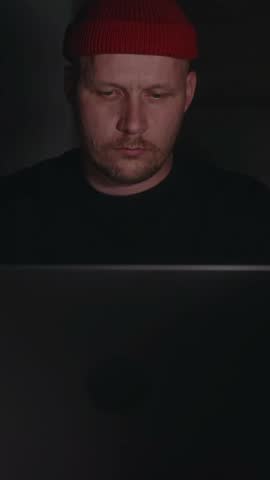 Vertical shot from behind the laptop showing a serious millennial man concentrating on his work in a dim room. Soft screen light highlights his focused expression and quiet nighttime workflow. 4k