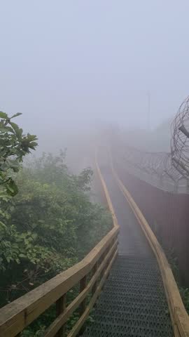 A dense fog envelops a wooden deck walkway next to a barbed wire fence. The mist creates a serene, mysterious atmosphere where nature and man-made structures merge.