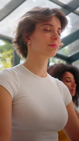 Close up of attractive caucasian female keeping eyes closed while meditating with diverse friends indoors. Attractive lady breathing slowly and enjoying calm atmosphere in spacious studio.