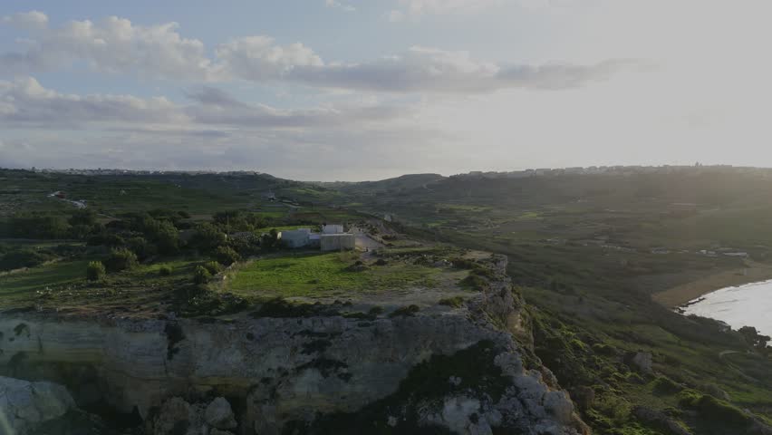 A smooth drone descent over Gozo reveals Mixta Cave glowing in warm sunset light, with rugged cliffs, blue sky, soft clouds and Mediterranean colors.