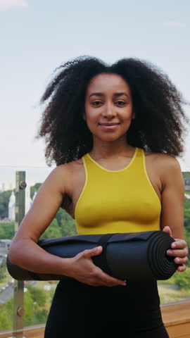 Slim African American sportswoman with rolled yoga mat stands on hotel rooftop. Sportive black lady in activewear radiates positiveness against urban park