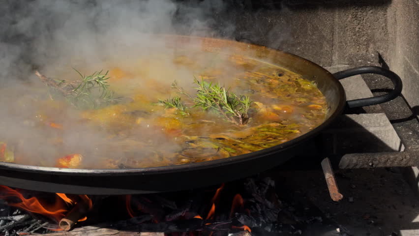 Cast-iron paella simmers over charcoal steam swirls around rosemary, evoking rustic, authentic Spanish outdoor cooking and warm tradition.