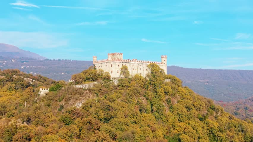 Aerial view of Castello di Montalto standing majestically on a hill surrounded by trees with autumn colors, under a clear blue sky, Castello di Montalto, Piemonte, italy.