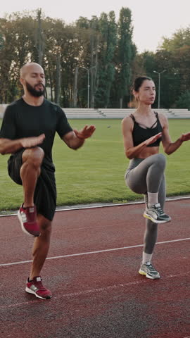Couple of athletic people works out on stadium in summer morning. Multinational man and woman train high knees running in place actively with palms above knees