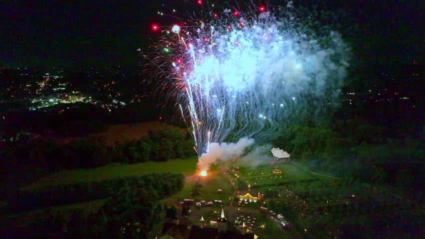 Bright fireworks burst in a colorful display across the night sky. Viewers enjoy the spectacle from a large gathering in a valley. The excitement creates a festive atmosphere.