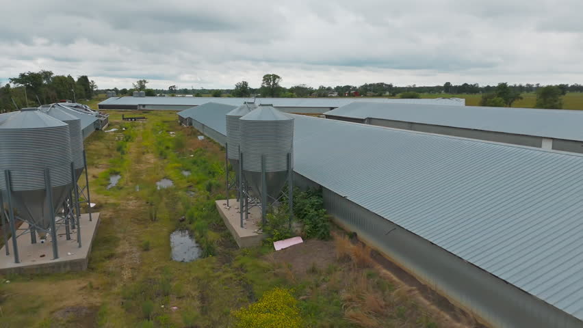 Farmland silos, metal industrial sheds, and water reservoir surrounded by trees under overcast sky, aerial establishing dolly between rows