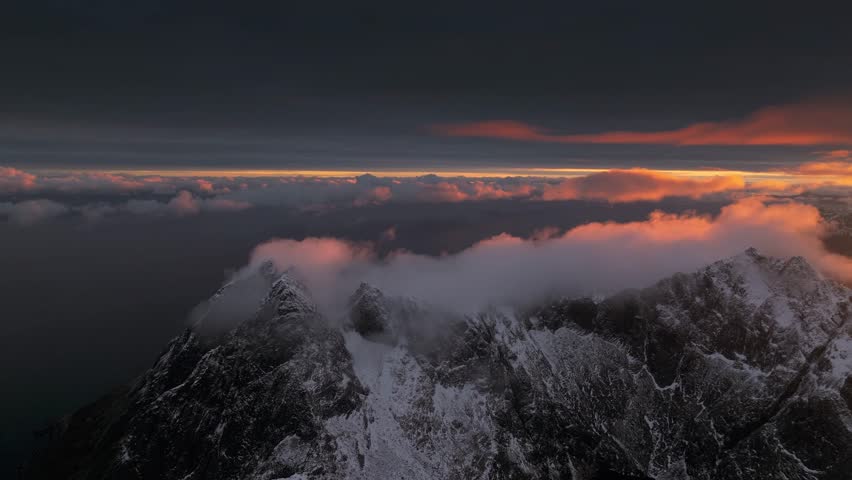 Aerial view of snow-covered mountain peaks kissed by the warm glow of the setting sun, creating a stunning contrast against the dark sky, Ballstad, Nordland, Norway.