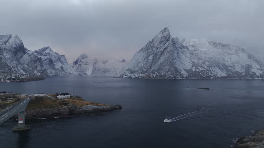 Aerial view of Reine's dramatic landscape, showcasing snow-covered mountains meeting the dark water, with a boat leaving a white trail, Reine, Nordland, Norway.