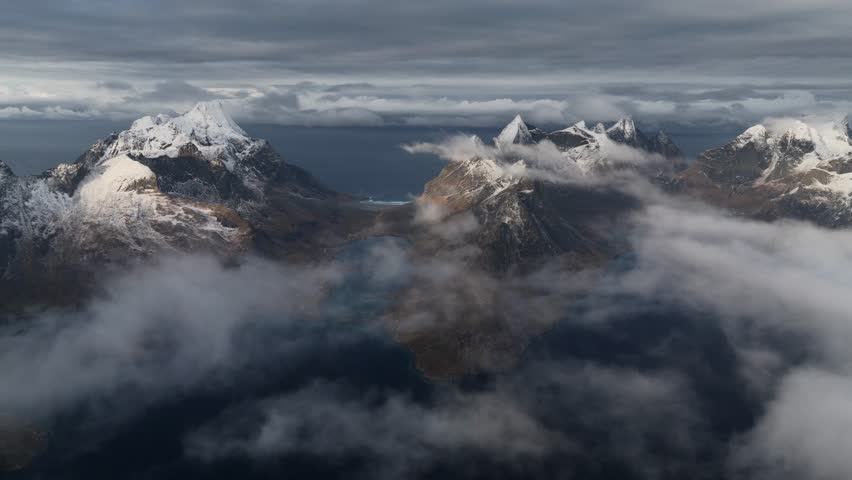 Aerial view of dramatic snow-capped peaks piercing through ethereal clouds, contrasting with the dark waters below, Reine, Nordland, Norway.