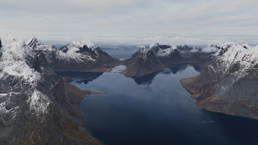 Aerial view of snow-capped mountains reflecting in the dark blue fjord waters, creating a striking contrast, Reine, Nordland, Norway.