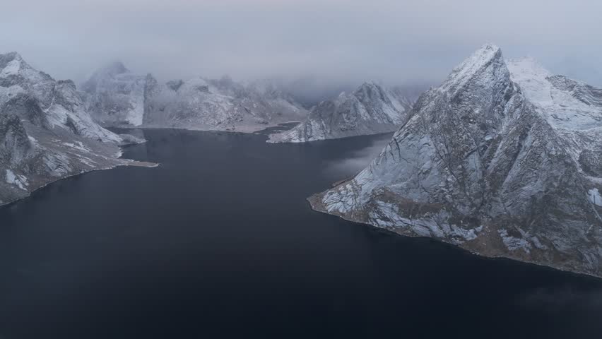 Aerial view of the snow-dusted mountain peaks, contrasting with the dark water below, creates a serene yet dramatic landscape, Reine, Nordland, Norway.