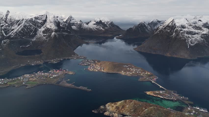 Aerial view of Reine, a fishing village beneath snow-dusted mountains and dark waters, linked by bridges, creating a striking contrast, Reine, Nordland, Norway.