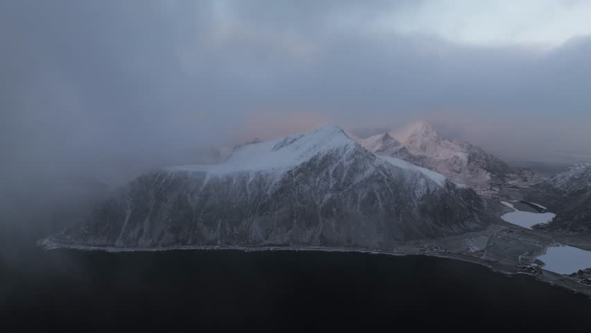 Aerial view of snow-capped mountain peaks shrouded in mist, creating a serene yet dramatic landscape with dark water and a cloudy sky, Reine, Nordland, Norway.