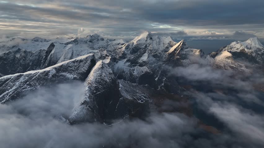Aerial view of snow-capped mountain peaks piercing through a sea of ethereal clouds, creating a stunning contrast of light and shadow, Reine, Nordland, Norway.