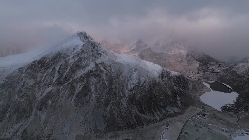 Aerial view of snow-capped mountains meet a serene lake under a cloudy sky, showcasing the stark beauty of the landscape, Reine, Nordland, Norway.
