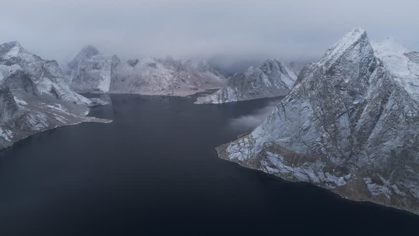 Aerial view of jagged, snow-dusted mountains meeting the still, dark lake under an overcast sky, creating stark contrast, Reine, Nordland, Norway.