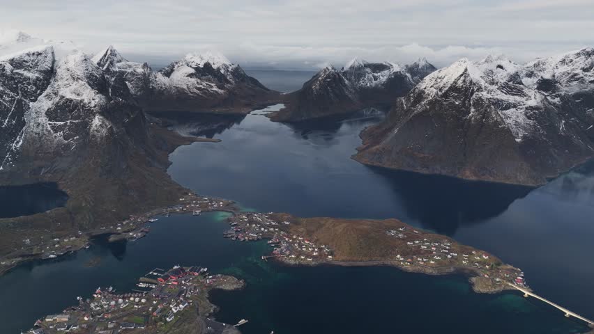Aerial view of Reine surrounded by dark blue water and snow-capped mountains under a cloudy sky, Reine, Nordland, Norway.