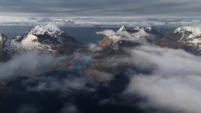Aerial view of snow-capped mountains piercing through a sea of clouds, creating a dramatic contrast of light and shadow, Reine, Nordland, Norway.