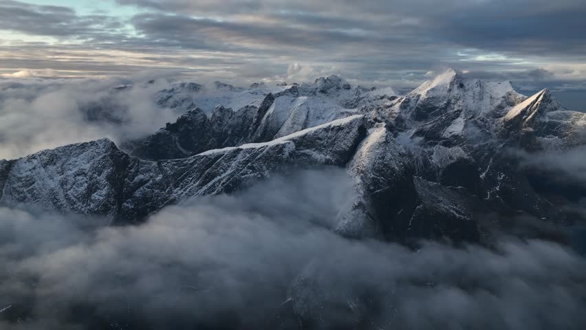 Aerial view of jagged, snow-dusted mountain peaks dramatically pierce through a sea of low-lying clouds, creating a serene yet powerful landscape, Reine, Nordland, Norway.