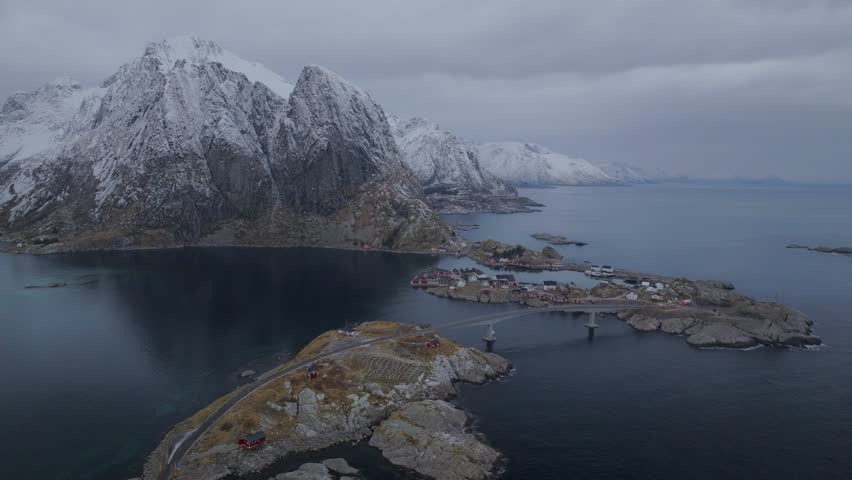 Aerial view of Reine, a fishing village, showcases red cabins and a bridge connecting rocky islands against a backdrop of snow-capped mountains, Reine, Nordland, Norway.