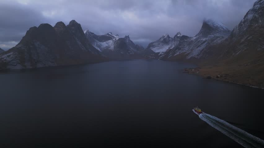 Aerial view of a boat sailing on dark waters near majestic, snow-capped mountains, under a cloudy sky, Reine, Nordland, Norway.