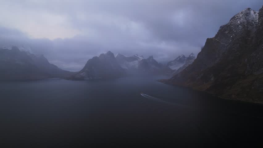 Aerial view of a boat sailing through the dark, still waters of a fjord surrounded by imposing, cloud-covered mountains, Reine, Nordland, Norway.