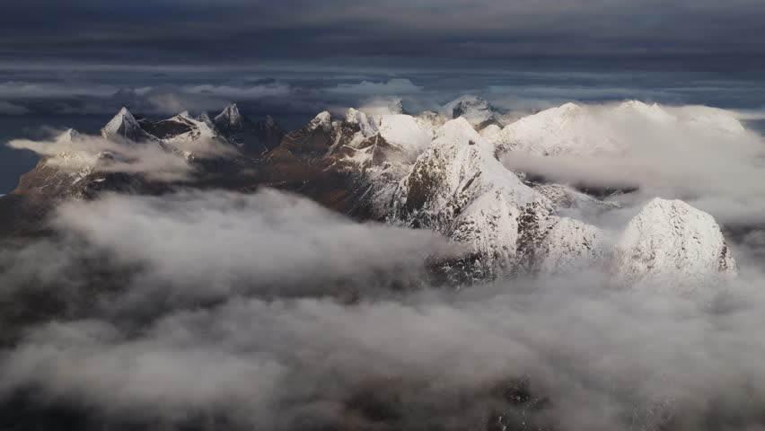 Aerial view of majestic snow-capped mountain peaks piercing through swirling clouds, a serene and dramatic landscape, Reine, Nordland, Norway.